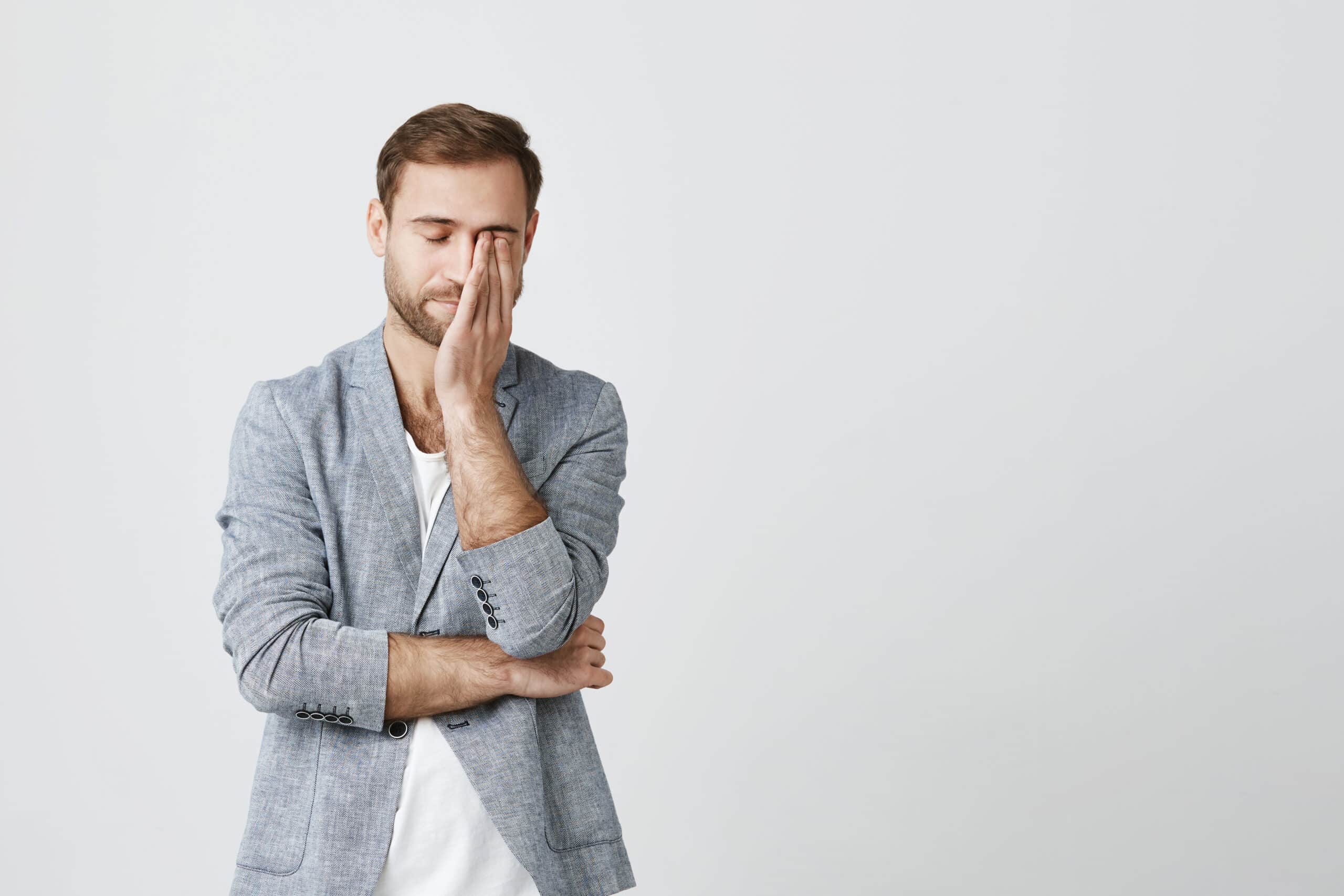 Attractive bearded male wears trendy clothes keeps hand on face, has headache after having conference, tired after hard working day, isolated against studio wall. Negative emotions, face expression